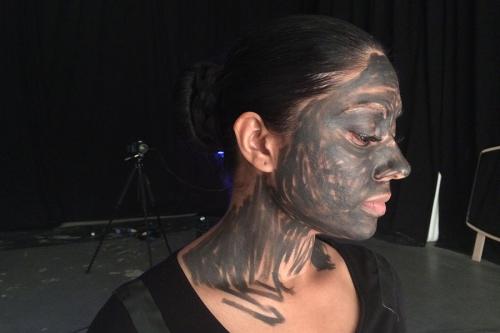 A woman in profile, with black hair tied back in a bun, black top, and black paint or makeup scribbled all over her face and neck.