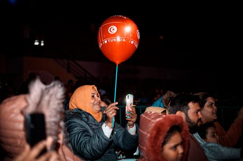 A young woman wearing a red jacket and black headscarf photographs a street procession on her phone.