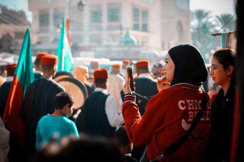 Amid a crowd, a woman wearing an orange headscarf films a performer while holding a red balloon with the Tunisian flag on it.