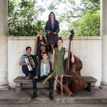 Five-piece band pose in a white marble structure, arranged in a pyramid shape and holding their instruments: drumsticks, violin, accordion, trumpet, and upright bass.