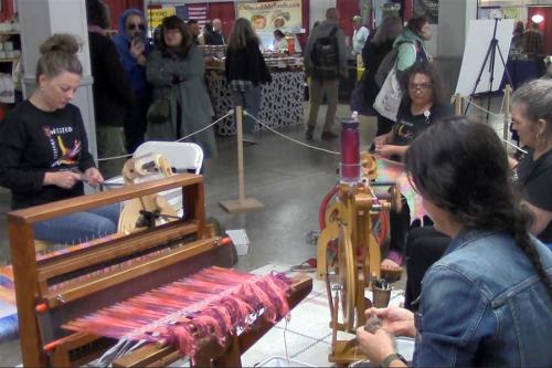 A small group in a circle works on yarn spinning wheels and a loom.