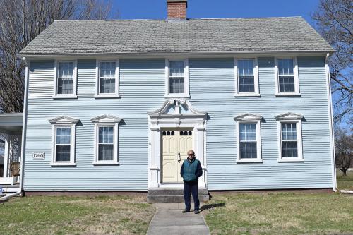 A man standing in front of a light blue two-story house with an ornate door frame and pale yellow door.