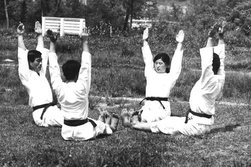 Four people wearing matching white martial arts uniforms with black belts sit in a circle on the ground, their legs straight forward and arms straight up. Black and white photo.