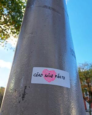 On a broad gray pole outdoors, a small white sticker with a pink heart reads Céad Míle Fáilte.