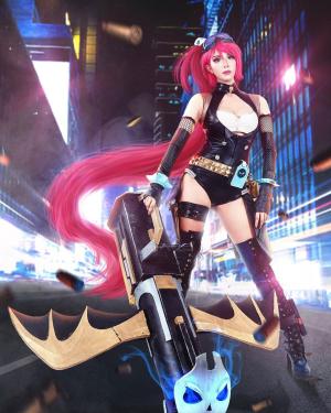 A person poses in a costume of very long windswept pink wig, goggles, black bodysuit, and tall black combat boots, holding some kind of rocket launcher. The costume is styled to look like a video game rendering, with a bright, blurry cityscape in the back