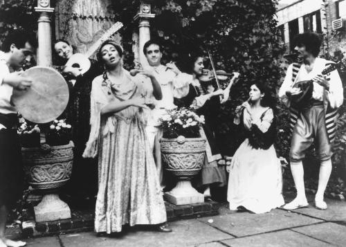 A group of people in old-fashioned clothes sing and play string instruments and hand drums in a courtyard.