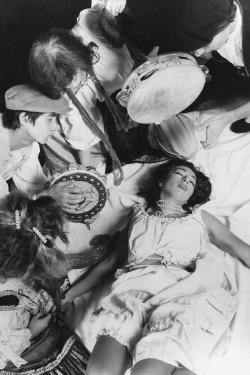 A woman in white dress lies on a white sheet on the ground, with drummers surrounding her.