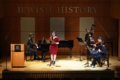 Musicians (pianist, vocalist, cellist, violinist, and clarinetist) on a stage under letters mounted on the back wall reading, Jewish History. 