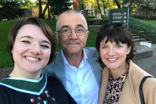Selfie of a young woman in a black graduation gown with multicolored embroidery in the front center, with her father and her mother, all smiling.