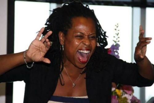 A Black woman acts out a part in a story, her hands raised as if pulling back curtains, her face menacing, like an animal about to attack.