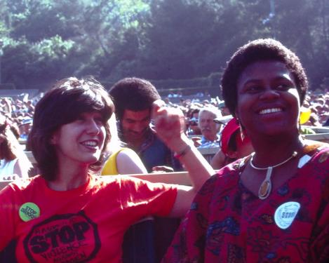 Two women smile among an audience in an outdoor amphitheater. One wears a bright red T-shirt that reads STOP violence against women.