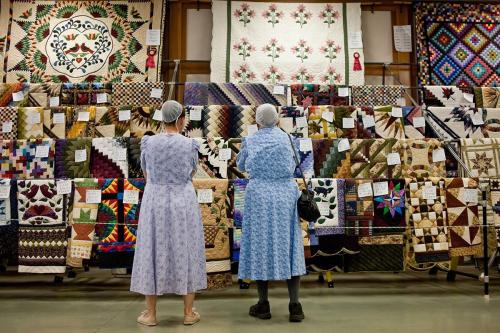 Two people, from behind, in dresses and cloth caps look at rows of hanging quilts.