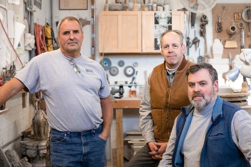 Three men post in a workshop, surrounding by toolds, cables, stone pieces, and cabinets.
