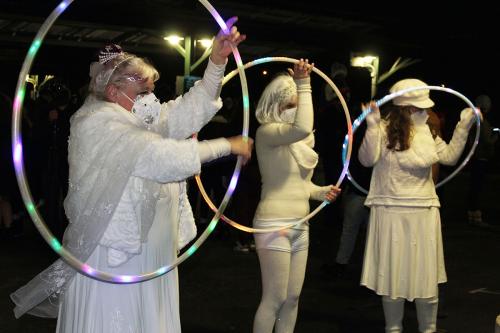 Three people dressed in all white - skirts, leggings, shirts, and face masks -  hold up lit-up Hula-Hoops.