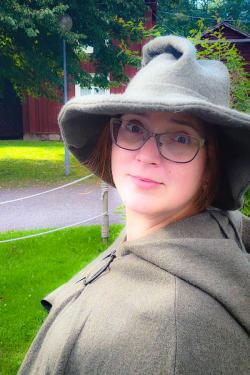 A person poses wearing a dark green-gray cloak and hat.
