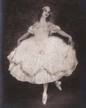 A ballerina poses in costume poses, arms outstreched, en pointe on one foot. Her white, tiered dress  billows out.