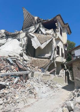 A three-story home is crumped, piles of rubble spilling out onto the dirt road.