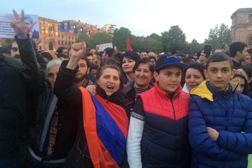 A crowd of people, including youth, facing forward. One woman in front raises her fist and wears an Armenian flag (red, blue, and orange stripes) like a bandolier. Color photo.
