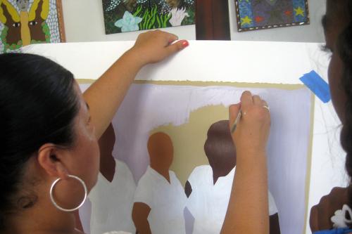 A person paints on a canvas, depicting dark-skinned figures. Their faces do not yet have any details. Another person watches the painter.