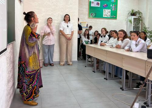A woman wearing large disc earrings and a colorful tunic speaks in front of a classroom of seated students in white dress clothes.