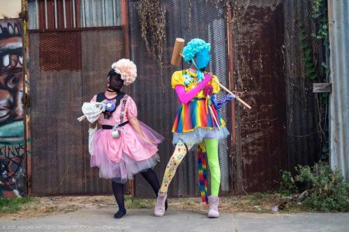 Two people pose in bright, multicolored dresses and poofy bonnets. One holds an oversized mallet. 