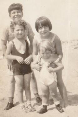 Four young kids pose on the beach in swimwear. One kid holds up the youngest of them, who looks down at the ground. Black-and-white photo.