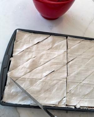 Closeup as someone cuts diagonally the unbaked, layered baklava with a knife.