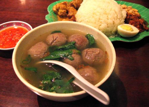 A bowl of brown beatballs and green leaves in a clear broth with a spoon. Behind it, a green platter with a big scoop of white rice, half a hard-boiled egg, peanuts, and brown meat.