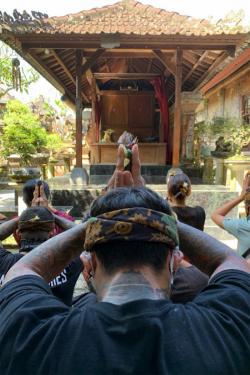 In the foreground is the back of someone’s head, head bowed and hands outstretched in prayer. In front of him are several other people in the same position, all facing a small outdoor temple with a tiled roof.