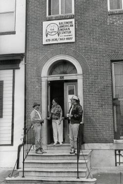 People gather on the front steps of a rowhouse building. A sign above the door reads: BALTIMORE AMERICAN INDIAN CENTER. Talk Together. With the address and phone number. Black-and-white photo.