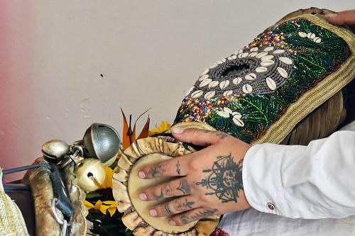 Close-up on a person playing a drum, which is adorned with a pattern of green sequins, gold trim, and shells. The one hand visible is tattooed with various symbols in black.
