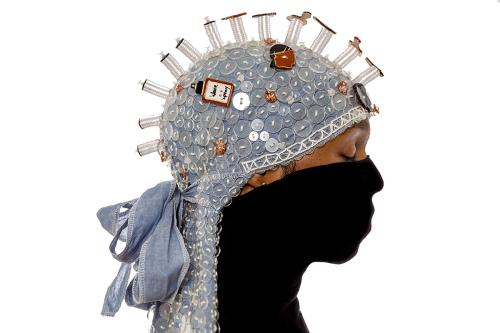 In profile, a person wearing a denim-colored durag around the dome of their head, draping down their neck. The fabric is covered in clear button, including stacks of buttons that appear like a row of spikes down the middle. Over their nose, mouth, and nec
