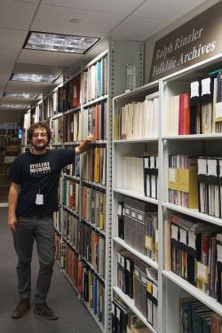 A young man poses along shelves full of records and binders. A sign on top of one shelf reads: Ralph Rinzler Folklife Archives