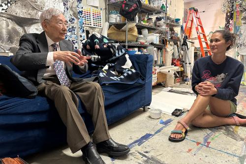 An elder man sits on a dark blue couch, speaking and gesticulating, while a young woman sitting cross-legged on the ground listens. Behind them are the clutters walls and shelves of a visual artist.