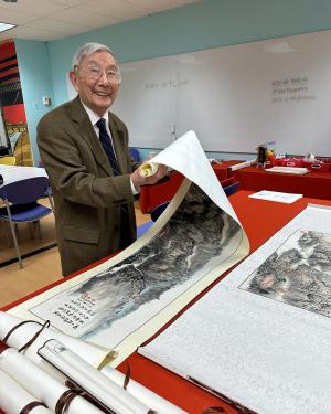 An elder man in a suit smiles as he rolls up a long painting on white paper. Other scrolls sit rolled up to the side.