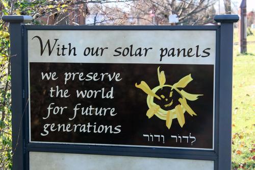 An outdoor sign reads: With our solar panels we preserve the world for future generations. On the right side, drawing of a yellow sun and Hebrew text.