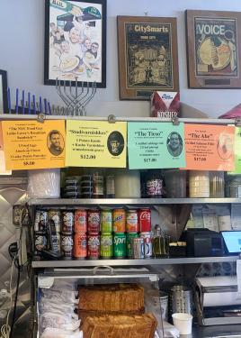 Behind the counter of a restaurant, with shelves of soda cans and sliced bread. Printed on bright colored paper, clipped along the top row of shelves, are a few of the restaurant's dishes: The NYC Food Guy Latke Lover's Breakfast Sandwich, The Tieso, The 