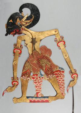 A two-dimensional, articulated shadow puppet of a human figure laying flat, with the rod to control it coming down from the hand on the right. Its body is painted gold with red embellishments, and its head is black, with facial features in gold and red.