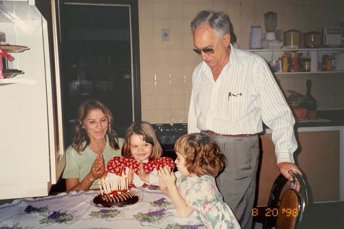 Two young girls and their grandmother sit at a table, with the grandfather standing beside them. A birthday cake with lit candles is on the table.