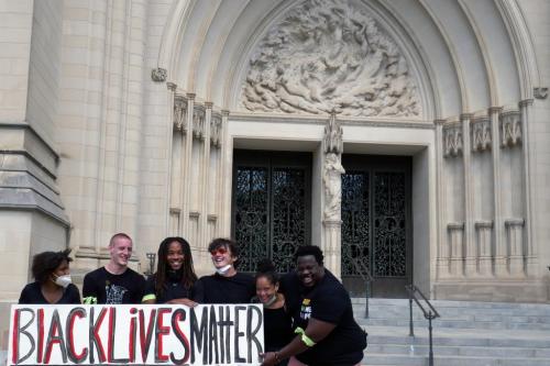 Six people—black, white, and smiling—hold up a protest sign that reads BLACK LIVES MATTER.