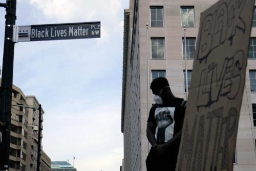 In the foreground, a cardboard sign reading BLACK LIVES MATTER. Behind it, a black man dressed in a black T-shirt showing a raised fist. To the side, a street sign for Black Lives Matter Plaza NW.