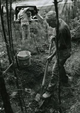 A man digs into the ground the a shovel. In the background, another person loads a bucket full of fresh clay into the back of a pickup truck. Black-and-white photo.