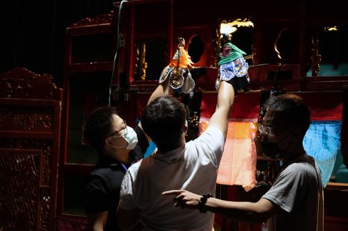 Three young men work the backstage of a puppet show. Two hold up glove puppets on their hands, while a third points somewhere offstage, perhaps cueing the band.