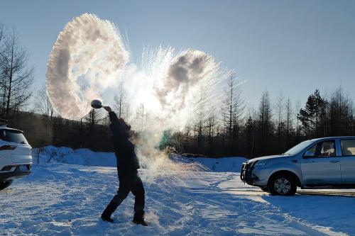 In silhouette, a person throw a pot of water into the air, which turns to streaks of white ice. The ground is covered in snow, and trees in the background are bare.