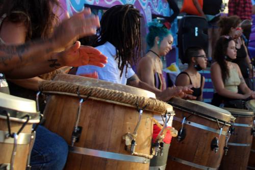 Close-up of four wooden barrel-shaped drums, held between the legs of their seated players.