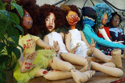Six cloth dolls lined up in sitting position. Each represents a woman with curly natural hair. One has a red hand shape painted over her mouth. Others have phrases written on their arms and feet.