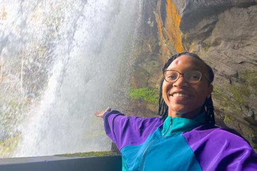 A young woman wearing glasses and purple and turquoise jacket smiles, gesturing behind her toward the backside of a waterfall along a rocky cliff.