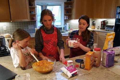 Three young people wearing aprons stand at a kitchen counter, with a mixing bowl of somethinng orange and lumpy and several bags, bottles, and boxes of ingredients in front of them. One person holds a glass mearuing container full of milk, looking nervous