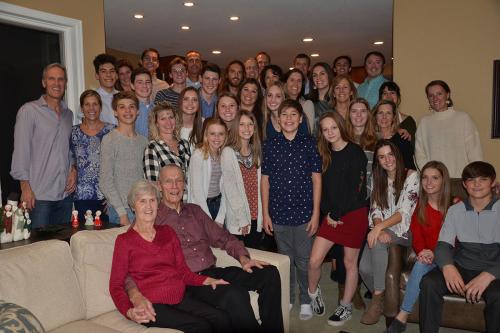 About 40 people, representing many generations of a family, pose in a living room, with two gransparents seated on a couch in front.