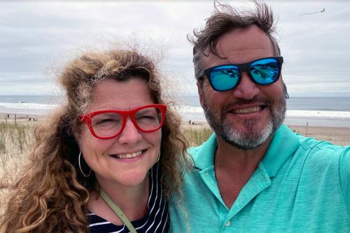 Woman in red glasses with long curly hair and man with blue reflective sunglasses pose together for a selfie in front of an ocean beach.